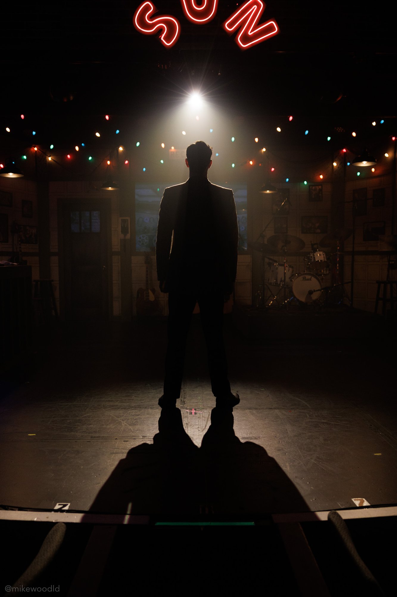 A silhouetted man stands center stage under a tight overhead special; a glowing red SUN sign and a string of multicolored holiday bulbs frame the bar set behind him